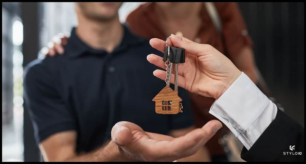 Un agente inmobiliario entrega un llavero con la forma de una casa a una pareja desenfocada en el fondo, simbolizando la compra de una nueva vivienda.