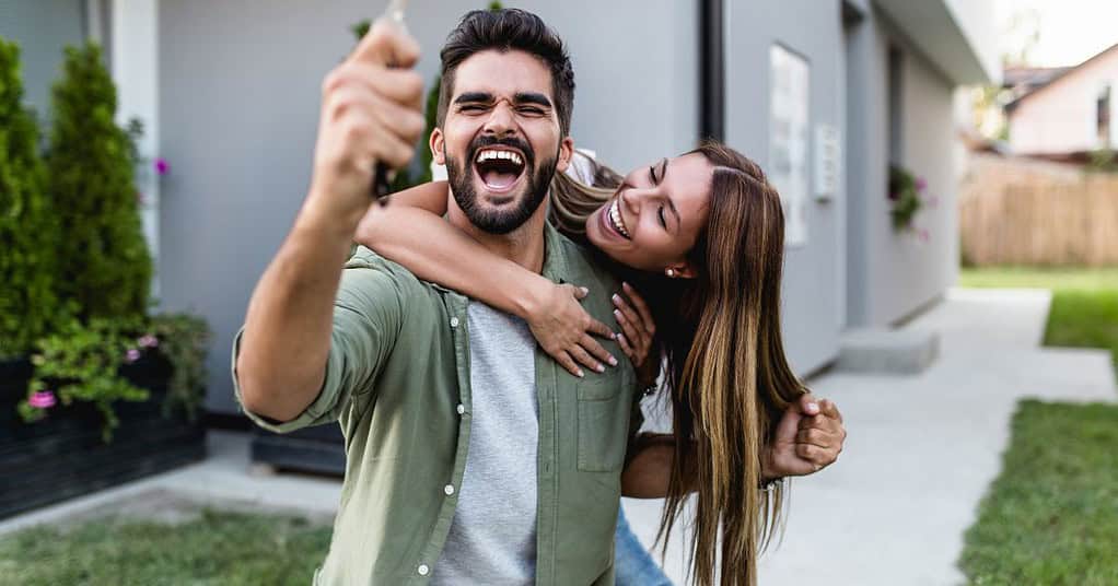 Una pareja joven celebra con entusiasmo fuera de su nueva casa, el hombre sostiene las llaves mientras la mujer lo abraza desde atrás. La imagen representa la alegría y la emoción de adquirir una propiedad.