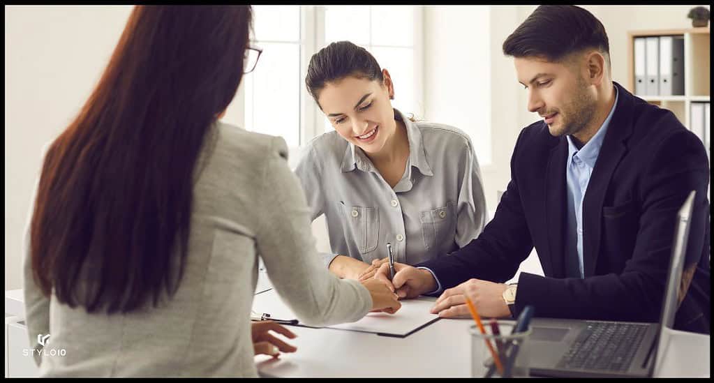 Una pareja sonriente firma documentos junto a una agente inmobiliaria en una oficina luminosa. Los tres están sentados alrededor de una mesa con una computadora portátil y papeles, simbolizando una transacción inmobiliaria