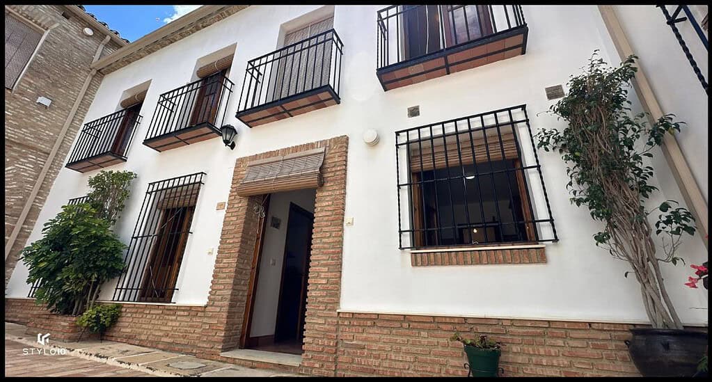 Fachada de una casa tradicional en Bujalance, con paredes blancas y detalles en ladrillo visto. Las ventanas y la puerta de entrada están protegidas por rejas de hierro negro, y la casa cuenta con pequeños balcones en las ventanas superiores. Una planta trepadora adorna la entrada, destacando el estilo andaluz de la vivienda.