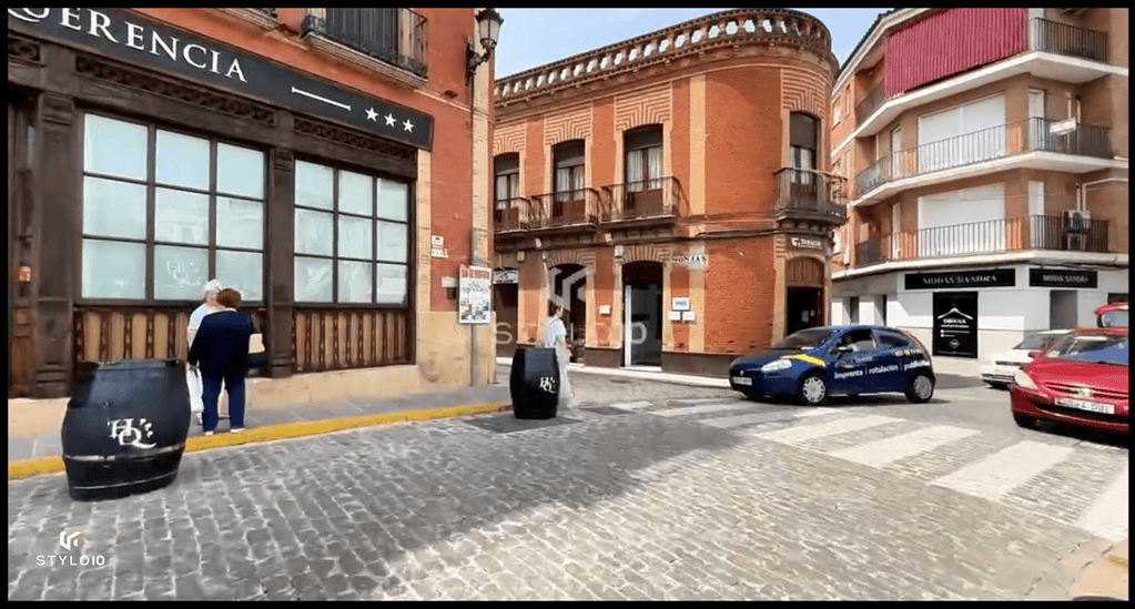 Calle empedrada en Bujalance, mostrando un edificio de ladrillo rojo con detalles arquitectónicos tradicionales y un hotel de cuatro estrellas. Se observan dos personas conversando en la acera, un coche azul publicitario y otros vehículos estacionados. La escena refleja la vida cotidiana en este pueblo andaluz.