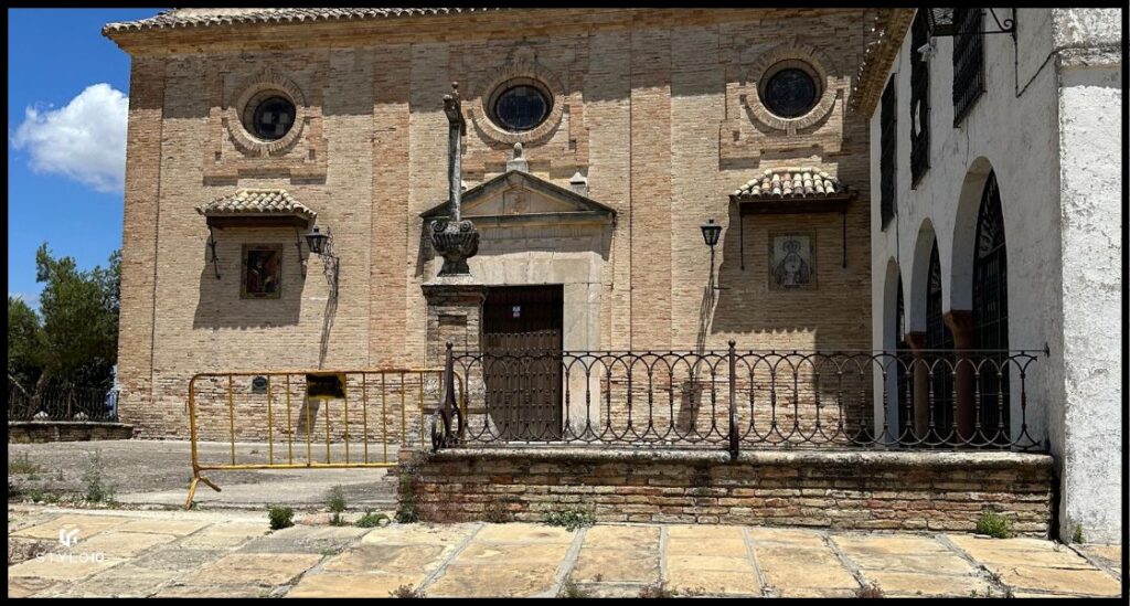 Fachada de la Ermita en Bujalance, construida en ladrillo visto con detalles arquitectónicos tradicionales. La entrada principal está flanqueada por una cruz de piedra y rodeada por una baranda de hierro. La imagen también muestra ventanales circulares y una pequeña capilla lateral, reflejando la arquitectura religiosa andaluza.