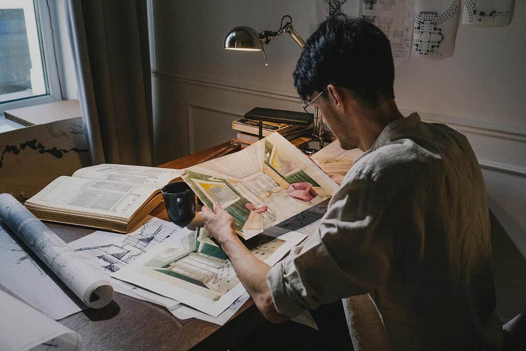 Architect focused on interior design sketches at desk with papers, lamp, and mug.